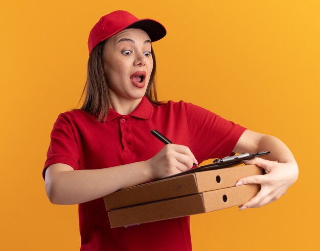 Entregadora bonita animada de uniforme escreve na prancheta com um marcador segurando em caixas de pizza isoladas na parede laranja com espaço de cópia