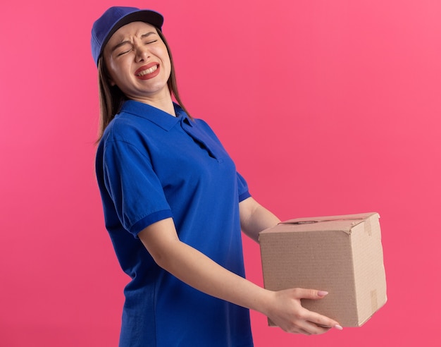 Entregadora bonita e insatisfeita de uniforme segura uma caixa de papelão isolada na parede rosa com espaço de cópia