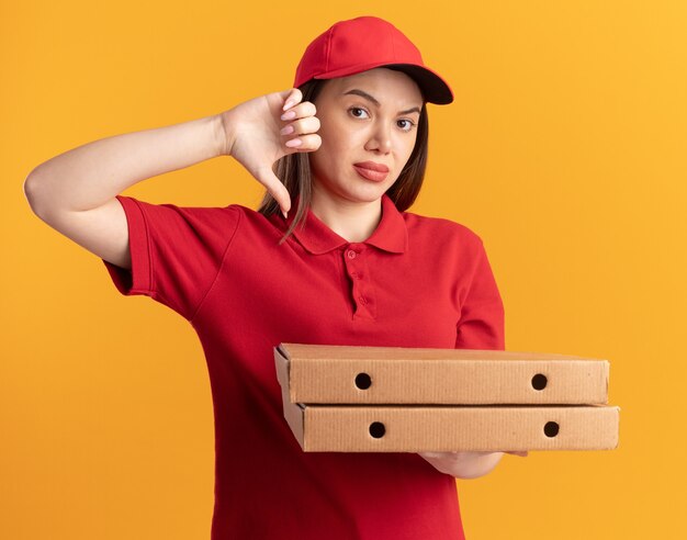 Entregadora bonita insatisfeita de uniforme com os polegares para baixo e segurando caixas de pizza em laranja