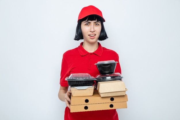 Entregadora caucasiana jovem e insatisfeita segurando recipientes de comida e embalagens em caixas de pizza e olhando