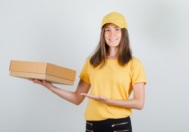 Entregadora de camiseta amarela, calça, boné mostrando caixa de papelão e parecendo alegre