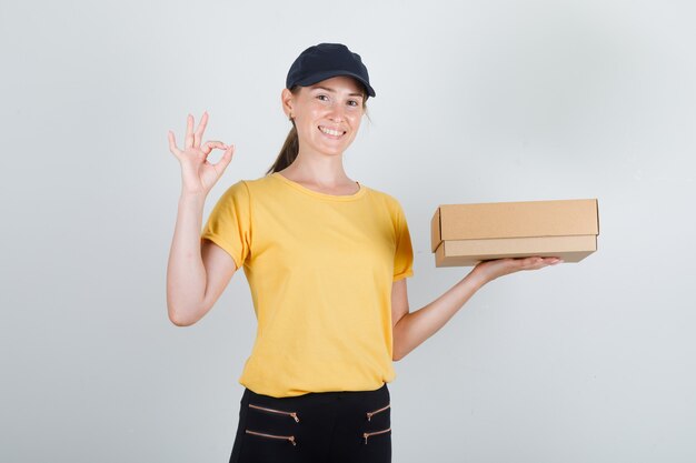 Entregadora de camiseta, calça e boné segurando uma caixa de papelão com o sinal de ok e parecendo satisfeita