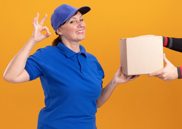 Entregadora de meia-idade com uniforme azul e boné dando uma caixa de papelão para um cliente sorrindo amigável mostrando uma placa de ok em cima da parede laranja