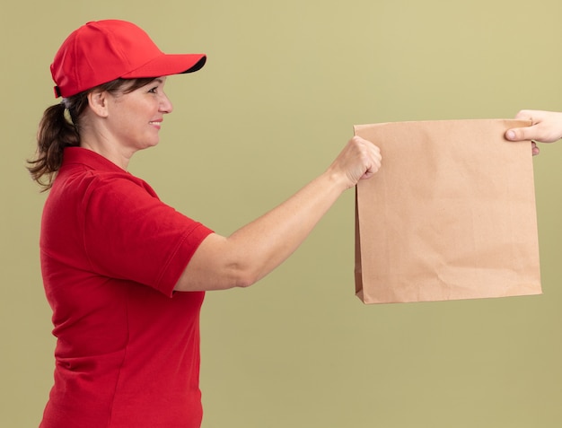 Foto grátis entregadora de meia-idade com uniforme vermelho e boné dando um pacote de papel para um cliente sorrindo amigável em pé sobre uma parede verde