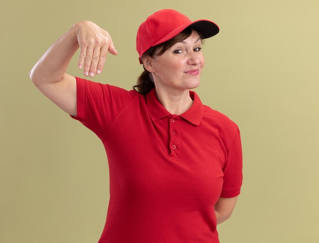 Entregadora de meia-idade com uniforme vermelho e boné gesticulando com as mãos parecendo confiante em pé sobre a parede verde