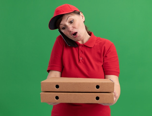 Entregadora de meia-idade com uniforme vermelho e boné segurando caixas de pizza parecendo confusa falando no celular em pé sobre a parede verde