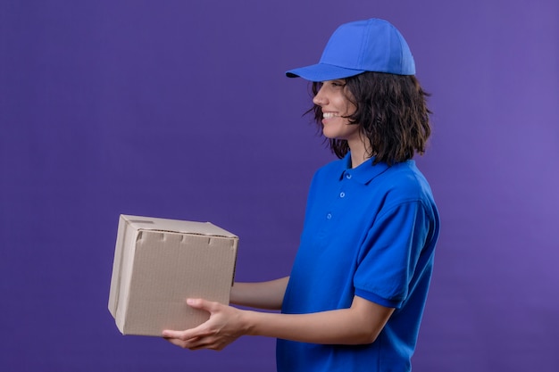 Entregadora de uniforme azul e boné de lado dando uma caixa para um cliente sorrindo amigável em um roxo isolado