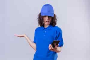 Foto grátis entregadora de uniforme azul e boné olhando para o celular com expressão confusa em pé com o braço levantado