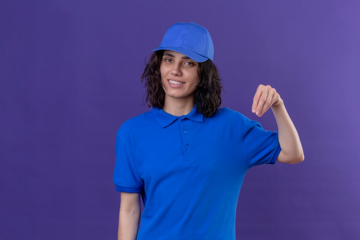 Foto grátis entregadora de uniforme azul e boné parecendo confiante gesticulando com a mão, conceito de linguagem corporal parado no roxo isolado