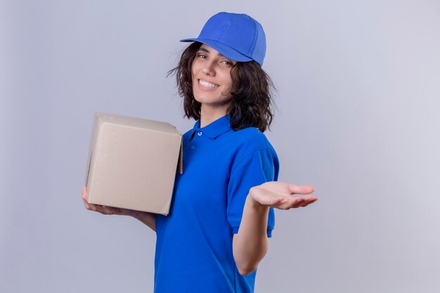 Entregadora de uniforme azul e boné segurando a embalagem da caixa fazendo um gesto de boas-vindas com a mão sorrindo amigável em pé no branco