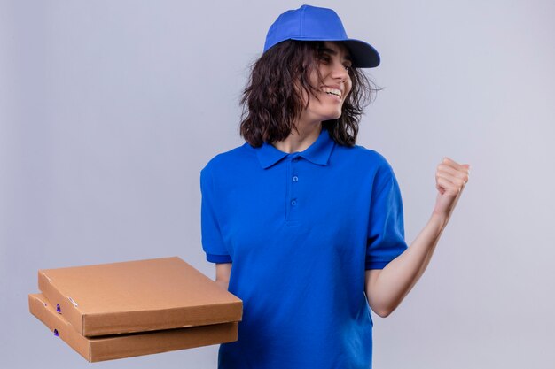 Entregadora de uniforme azul e boné segurando caixas de pizza apontando para trás com a mão e o polegar sorrindo alegremente em pé no branco