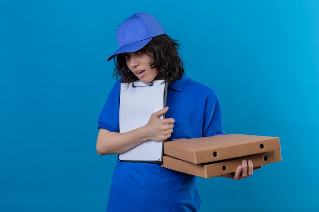 Foto grátis entregadora de uniforme azul e boné segurando caixas de pizza e prancheta pedindo assinatura, parecendo trabalhar em pé no azul isolado