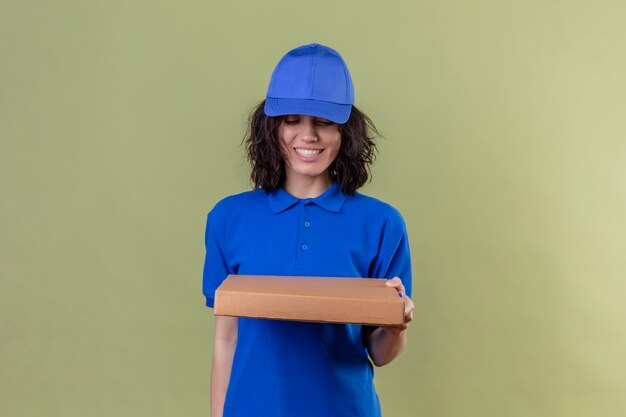 Foto grátis entregadora de uniforme azul e boné segurando uma caixa de pizza sorrindo alegre olhando para a caixa em pé sobre o espaço de cor verde-oliva isolado