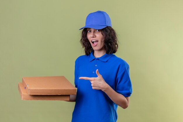 Entregadora de uniforme azul segurando caixas de pizza e apontando com o dedo indicador para elas sorrindo com uma carinha feliz em pé sobre o espaço verde