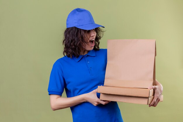 Entregadora de uniforme azul segurando caixas de pizza e um pacote de papel parecendo surpresa e feliz em pé no espaço verde