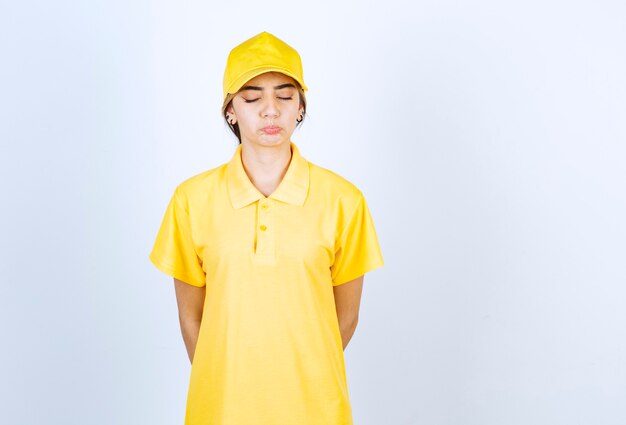 Entregadora em uniforme amarelo em pé com os olhos fechados contra uma parede branca.