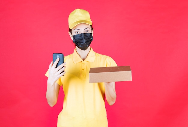 Entregadora feminina em uniforme amarelo e máscara preta segurando a caixa de papelão e fazendo uma videochamada ou recebendo pedidos via smartphone, parecendo confusa