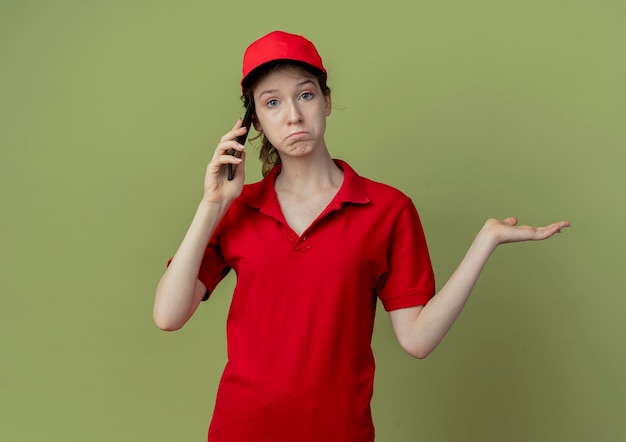 Entregadora jovem bonita confusa de uniforme vermelho e boné, falando no telefone e mostrando a mão vazia, isolada em um fundo verde oliva com espaço de cópia