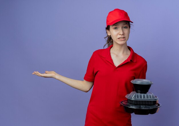 Entregadora jovem bonita descontente vestindo uniforme vermelho e boné segurando recipientes de comida e mostrando a mão vazia isolada em um fundo roxo com espaço de cópia