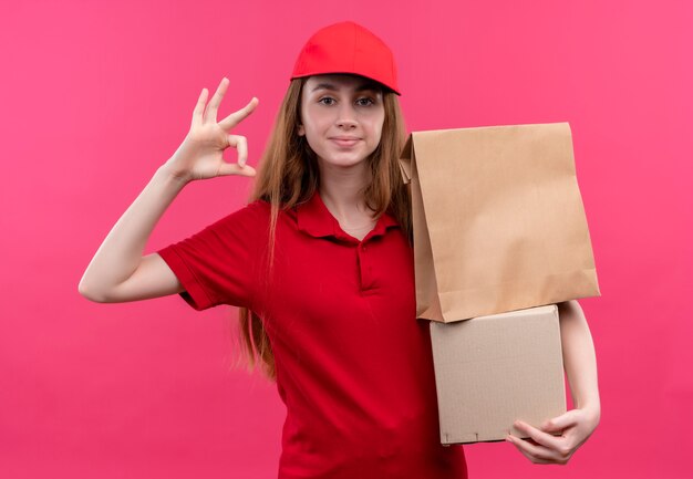 Entregadora jovem confiante em uniforme vermelho segurando caixas e fazendo sinal de ok no espaço rosa isolado