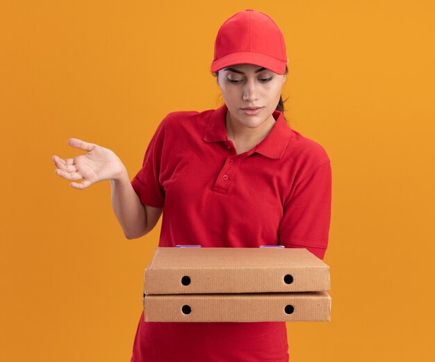 Entregadora jovem confusa vestindo uniforme e boné segurando e olhando para caixas de pizza espalhando a mão isolada na parede laranja