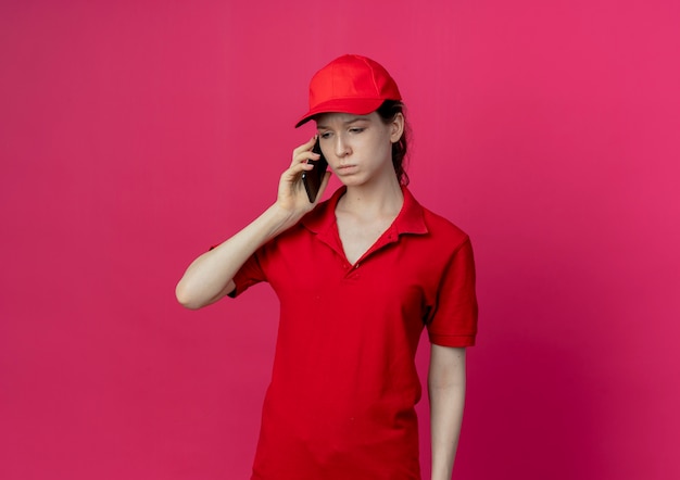 Entregadora jovem e desagradável com uniforme vermelho e boné falando no telefone, olhando para baixo, isolada no fundo carmesim com espaço de cópia