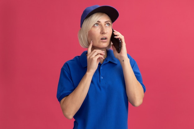 Entregadora loira de meia-idade impressionada com uniforme azul e boné falando ao telefone, olhando para cima tocando o rosto com o dedo