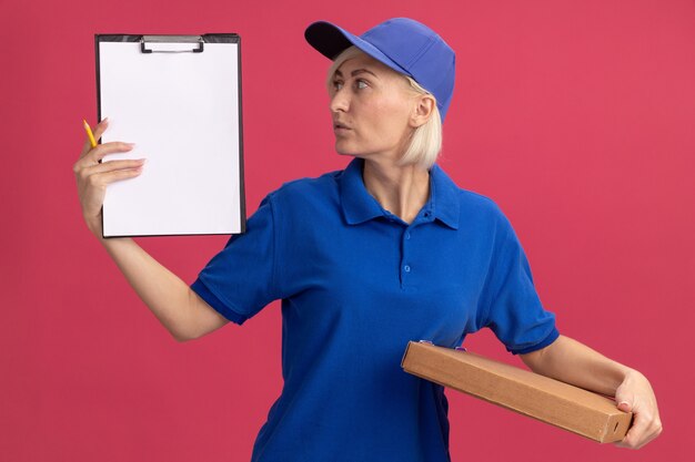 Entregadora loira de meia-idade impressionada com uniforme azul e boné segurando um pacote de pizza de lápis para prancheta, olhando para a prancheta isolada na parede rosa