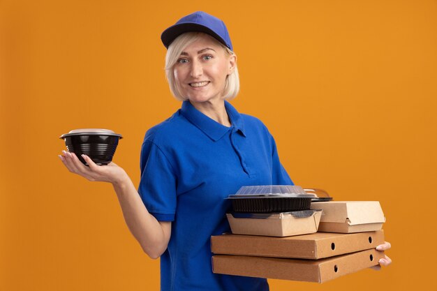 Entregadora loira de meia-idade sorridente com uniforme azul e boné segurando pacotes de pizza com recipientes de comida e um pacote de comida de papel neles isolado na parede laranja