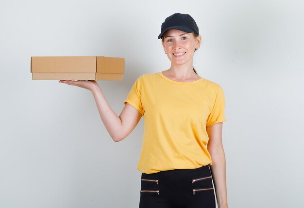Entregadora segurando uma caixa de papelão e sorrindo com camiseta, calça e boné