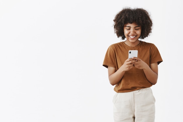 Foto grátis entretido fofa feliz adolescente afro-americana com penteado afro em uma camiseta marrom segurando um smartphone e rindo de um vídeo engraçado na internet usando o dispositivo para se divertir