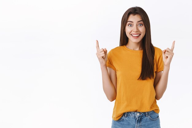 Entusiasmada, impressionada e animada sorrindo, mulher morena feliz em t-shirt amarela, rindo e sorrindo emocionada, mal posso esperar para experimentar uma proposta de venda incrível, apontando para um anúncio legal