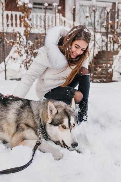 Entusiasta dama europeia olha com amor para seu cachorro, cansado após o jogo na neve. Menina bonita em um elegante traje branco brincando com husky em um dia gelado.