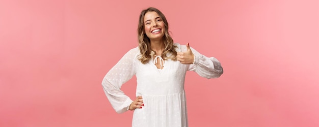 Foto grátis entusiasta e adorável jovem de vestido branco bonito mostra o polegar para cima em aprovação recomendo produto sorrindo satisfeito deixar impressão de revisão positiva em pé fundo rosa