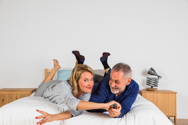 Foto grátis envelhecido mulher parando sorridente homem com controle remoto da tv para mudar de canal na tv na cama