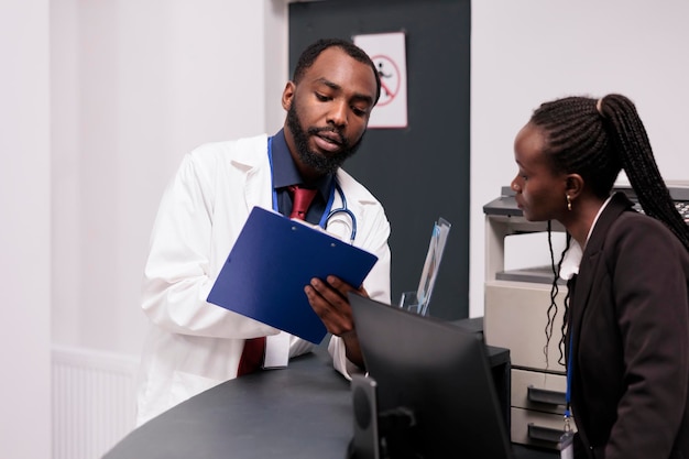 Equipe afro-americana trabalhando em relatórios de check-up na recepção do hospital, analisando consultas de pacientes em papéis. Médico e recepcionista usando formulários médicos para ajudar pessoas com doenças.