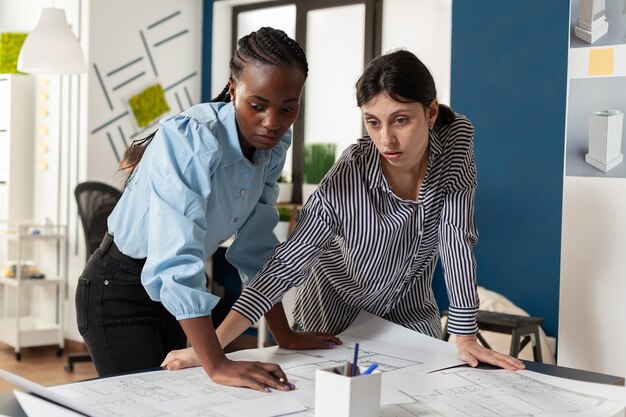 Equipe comprometida de parceiros de engenheiros discutindo planos de construção em pé na mesa do escritório de arquitetura moderno. Equipe de duas mulheres arquitetas projetando plantas para projeto de habitação imobiliária.