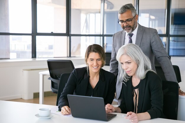 Equipe de negócios alegre assistindo apresentação no laptop, sentado no local de trabalho, olhando para o visor e sorrindo. Copie o espaço. Conceito de reunião de negócios