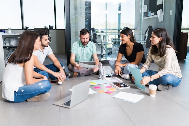 Equipe de negócios jovem sentado com o gerente no chão enquanto discutia no escritório criativo durante a reunião