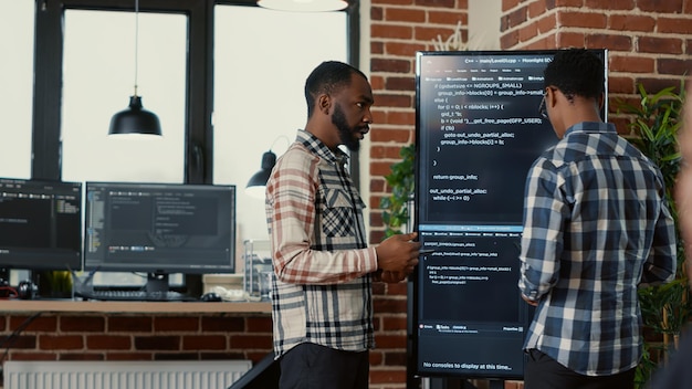 Equipe de programadores de software saindo depois de analisar o código-fonte na tv de tela de parede comparando erros usando tablet digital. Engenheiros de sistema passando telas compilando código para inteligência artificial.