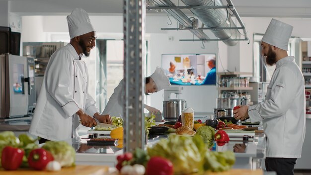 Equipe diversificada de chefs fazendo preparações de refeições com ingredientes, cozinhando deliciosos pratos gourmet na cozinha do restaurante. Pessoas de uniforme trabalhando como cozinheiros preparando receita de comida de gastronomia.