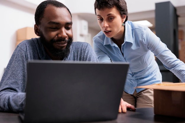 Equipe diversificada sentada à mesa no depósito verificando a logística de remessa no laptop antes de começar a entregar roupas da moda para os clientes. Funcionários que trabalham com caixas de papelão no armazém de moda