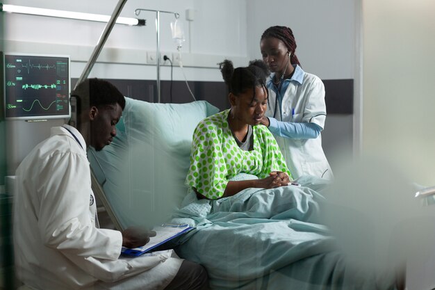 Equipe médica afro-americana que trabalha na cura de um paciente na cama da enfermaria do hospital. Homem e mulher com ocupação médica, examinando o jovem adulto para tratamento, usando monitor e estetoscópio