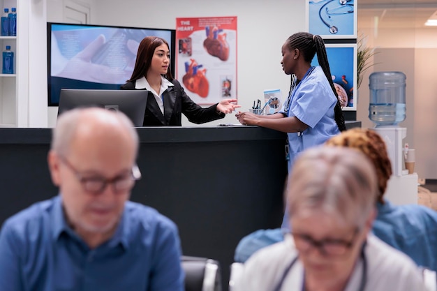 Equipe médica diversificada trabalhando no balcão da recepção, preenchendo papéis de relatório de check-up e marcando consultas. Enfermeira e recepcionista fazendo trabalho de registro na sala de espera do hospital.