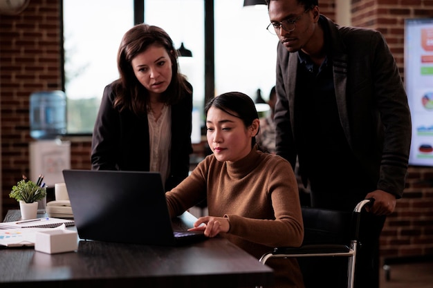 Equipe multiétnica de parceiros trabalhando com mulher paralisada sentada em cadeira de rodas, sofrendo de deficiência crônica e deficiência. Pessoas de negócios usando laptop, condição de saúde.