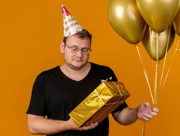 Eslavo adulto desapontado com óculos óticos e boné de aniversário segurando balões de hélio e uma caixa de presente