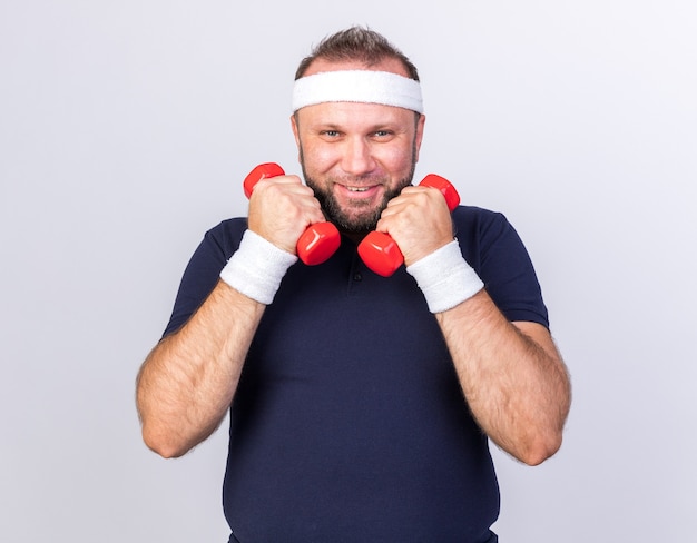 Foto grátis eslavo adulto e esportivo sorridente, usando bandana e pulseiras segurando halteres isolados na parede branca com espaço de cópia