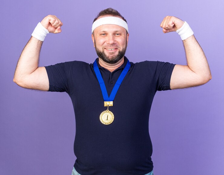 Foto grátis eslavo adulto sorridente homem desportivo com medalha de ouro em volta do pescoço, usando bandana e pulseiras tensionando os bíceps isolados na parede roxa com espaço de cópia