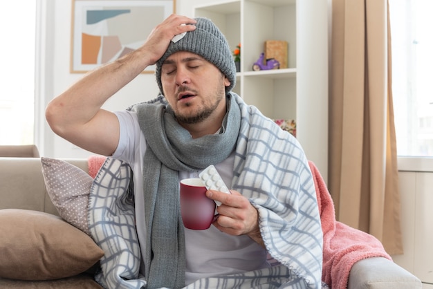 Eslavo doente e dolorido com um lenço no pescoço, um chapéu de inverno, colocando a mão na cabeça e segurando um copo com pacotes de remédios, sentado no sofá da sala