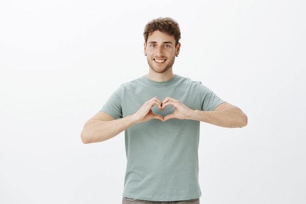 Espalhe o amor, não a guerra. retrato de um cara feliz de aparência amigável e positivo em brincos mostrando um gesto de coração sobre o peito e sorrindo amplamente enquanto expressa afeto ou emoções românticas sobre uma parede cinza